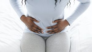 Afro woman holding belly, sitting on bed at home