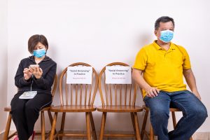 People with face mask seated in-between two chairs with Social Distancing, Do Not Sit signage.